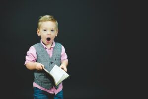 A boy wearing a gray vest and pink dress shirt holding a book